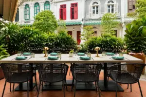 Outdoor restaurant terrace with a long dining table, green place settings, lush plants, and colorful heritage buildings.