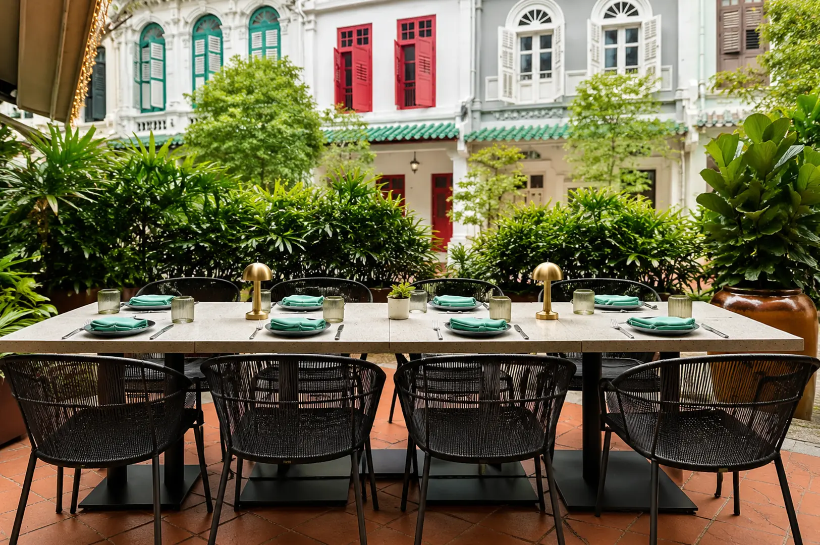 Outdoor restaurant terrace with a long dining table, green place settings, lush plants, and colorful heritage buildings.