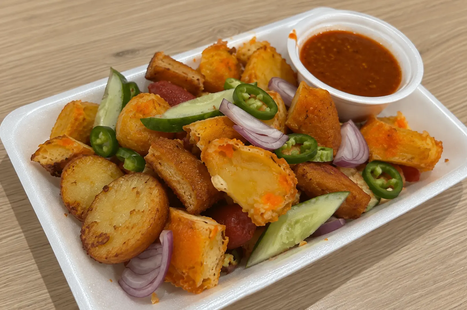 A tray of assorted fried snacks including fish cake, fried pieces, cucumber, chilies, onions, and dipping sauce.