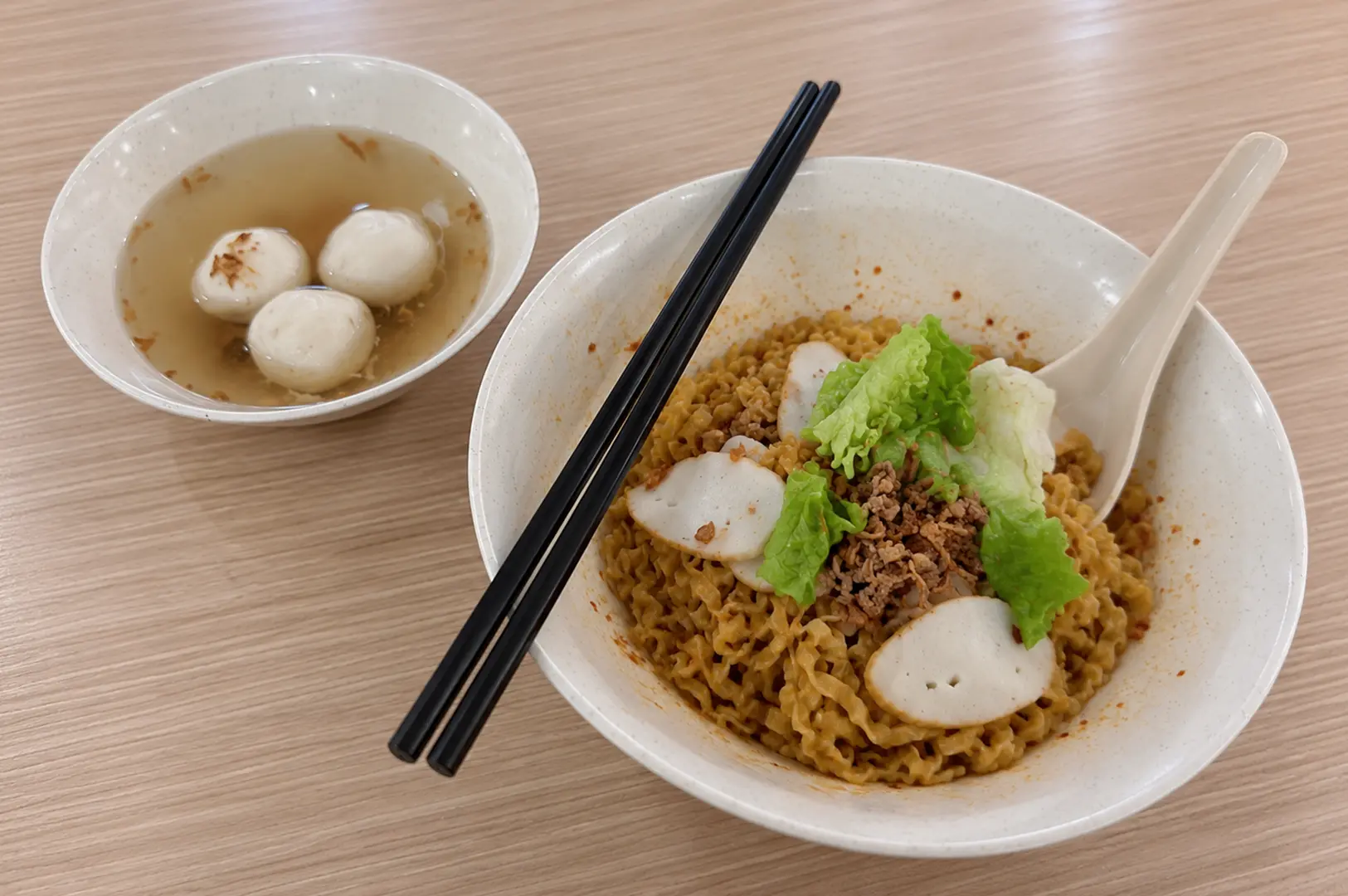 A bowl of dry mee pok topped with fish cake, minced meat, and lettuce, served with a separate bowl of fishball soup.