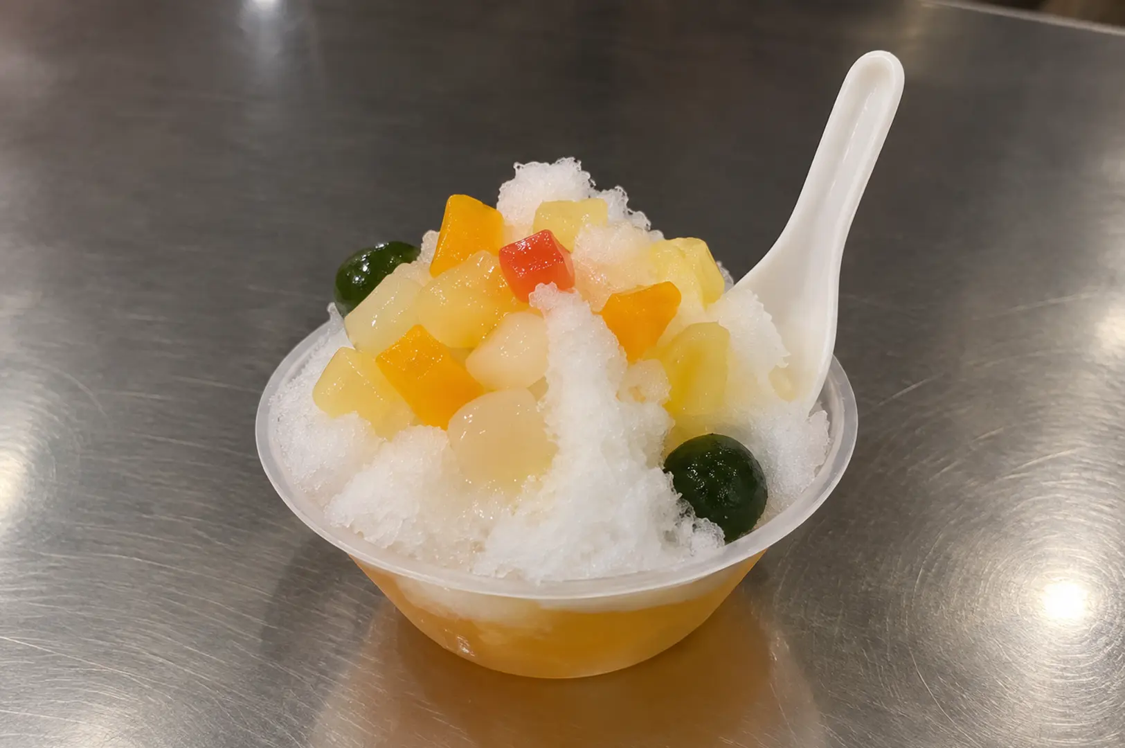 A chilled dessert topped with shaved ice, colorful fruit pieces, and sweet toppings in a clear bowl.