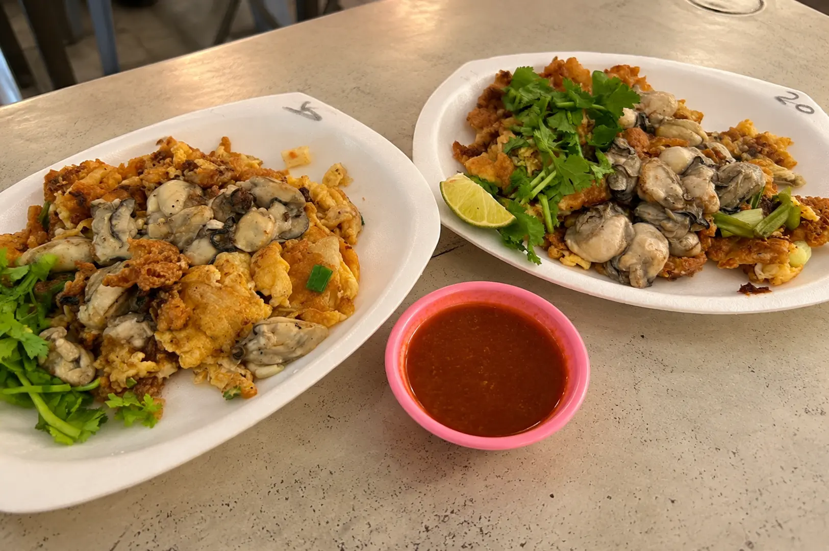 Two plates of Hup Kee oyster omelette topped with fresh oysters, crispy egg batter, coriander, lime, and a side of dipping sauce