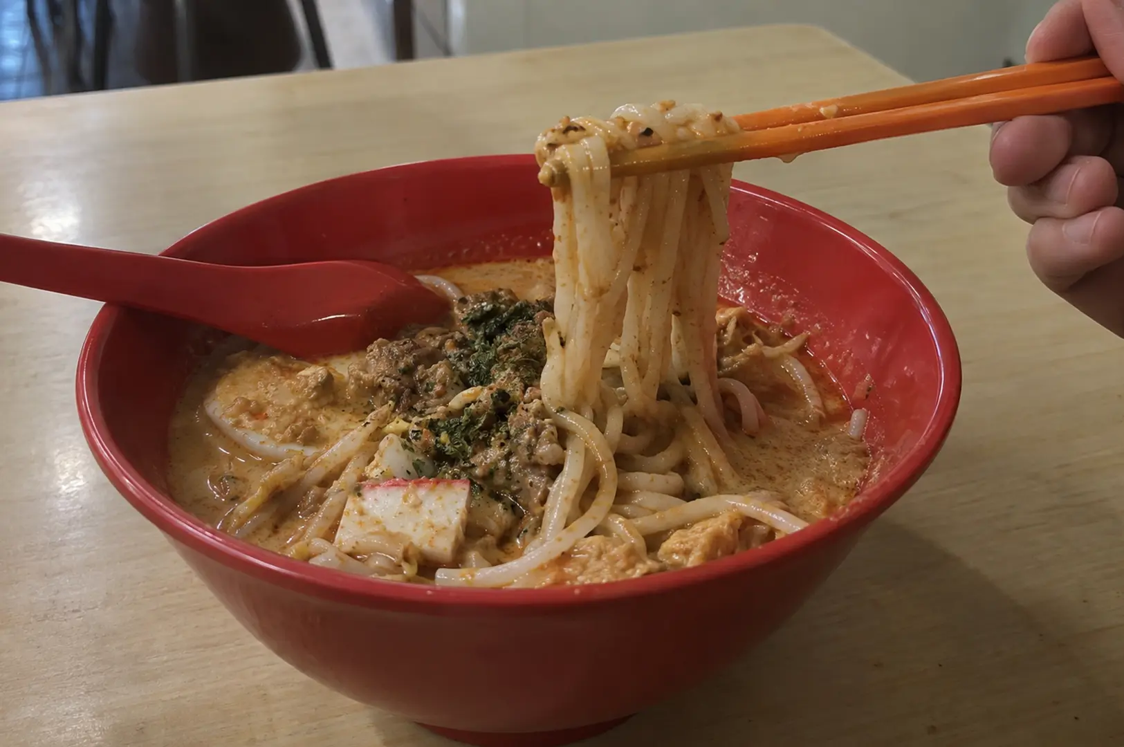 A bowl of creamy laksa with noodles lifted by chopsticks, showing tofu, toppings, and rich broth.