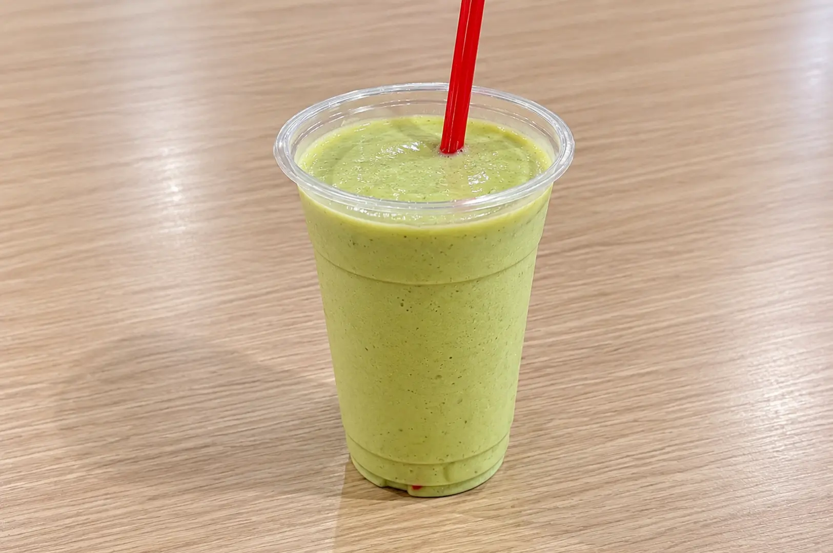 A creamy green avocado drink served in a clear plastic cup with a red straw on a wooden table.