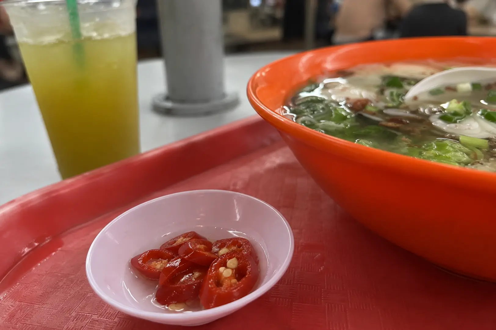 Bowl of fish porridge with leafy greens beside sliced red chilies and a refreshing drink.