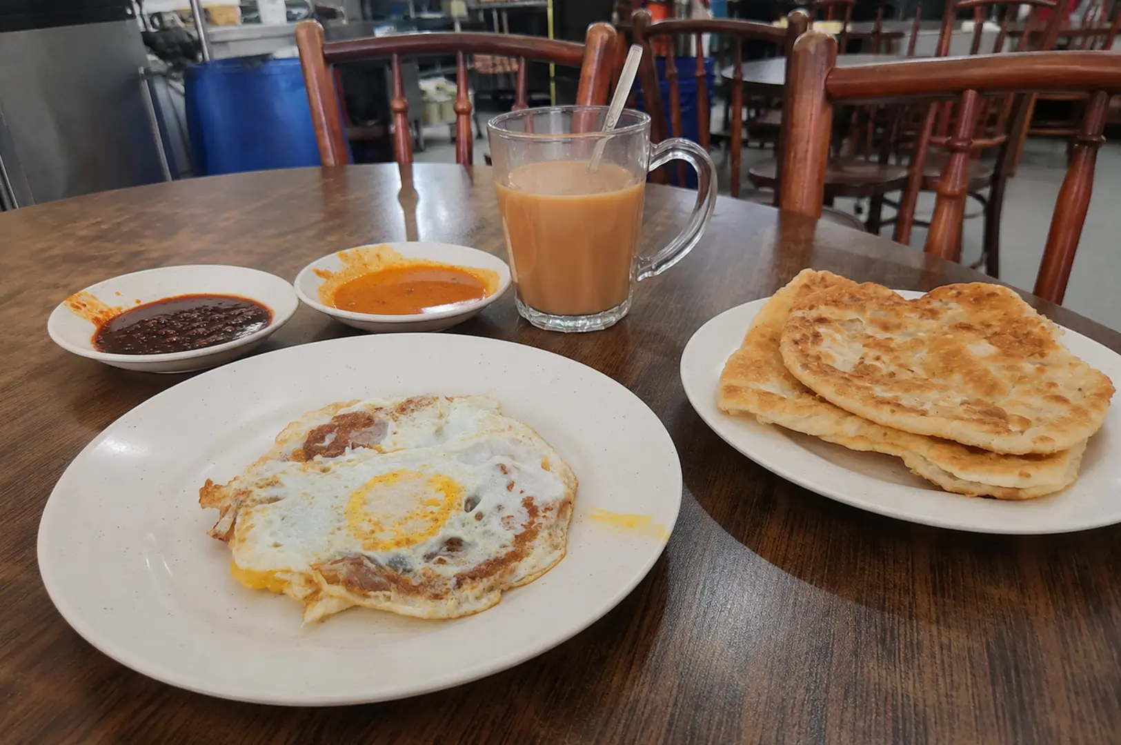 Roti canai with fried egg, curry dipping sauces, and a glass of hot milk tea.