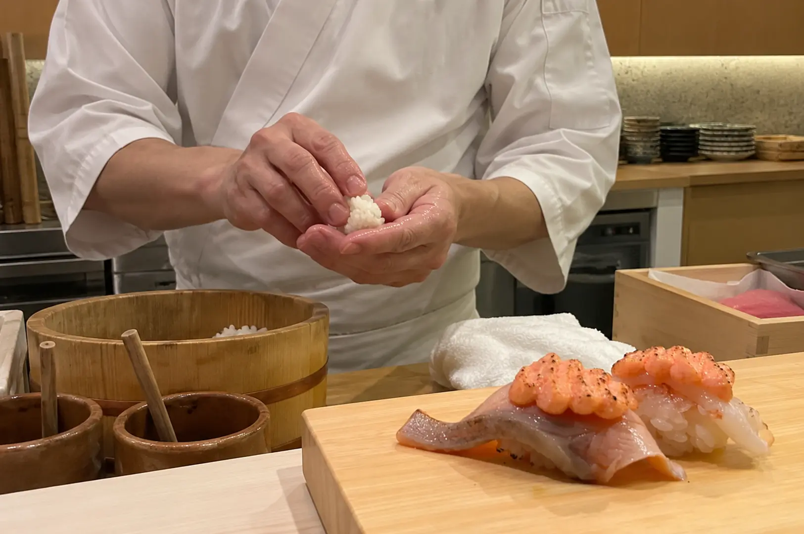 Sushi chef shaping rice by hand behind a counter with freshly prepared nigiri in the foreground.