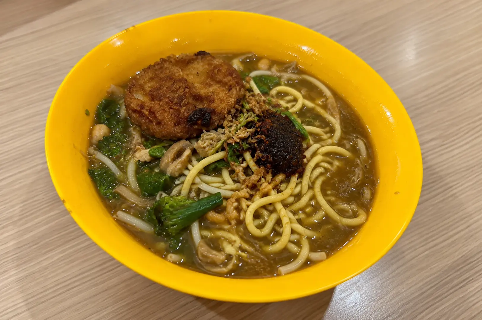 A bowl of mee rebus with yellow noodles, rich gravy, vegetables, fried toppings, and a crispy patty.