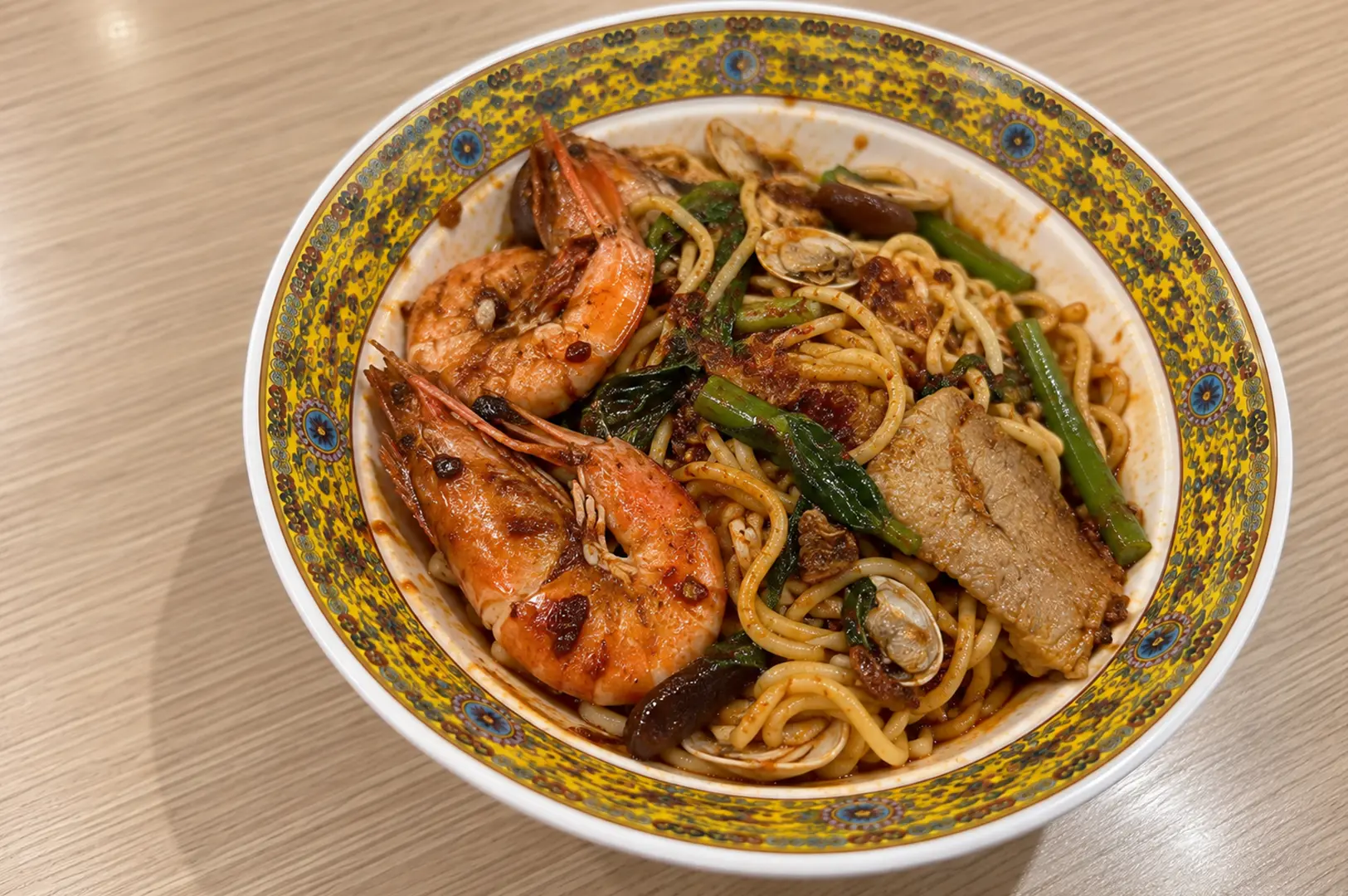 A bowl of pork ribs prawn noodles topped with large prawns, pork, clams, vegetables, and noodles.