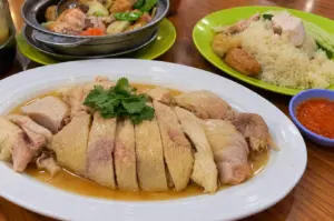 A plate of sliced steamed chicken with fragrant sauce, fresh coriander, red chilli dip, and a plate of chicken rice on a wooden table.