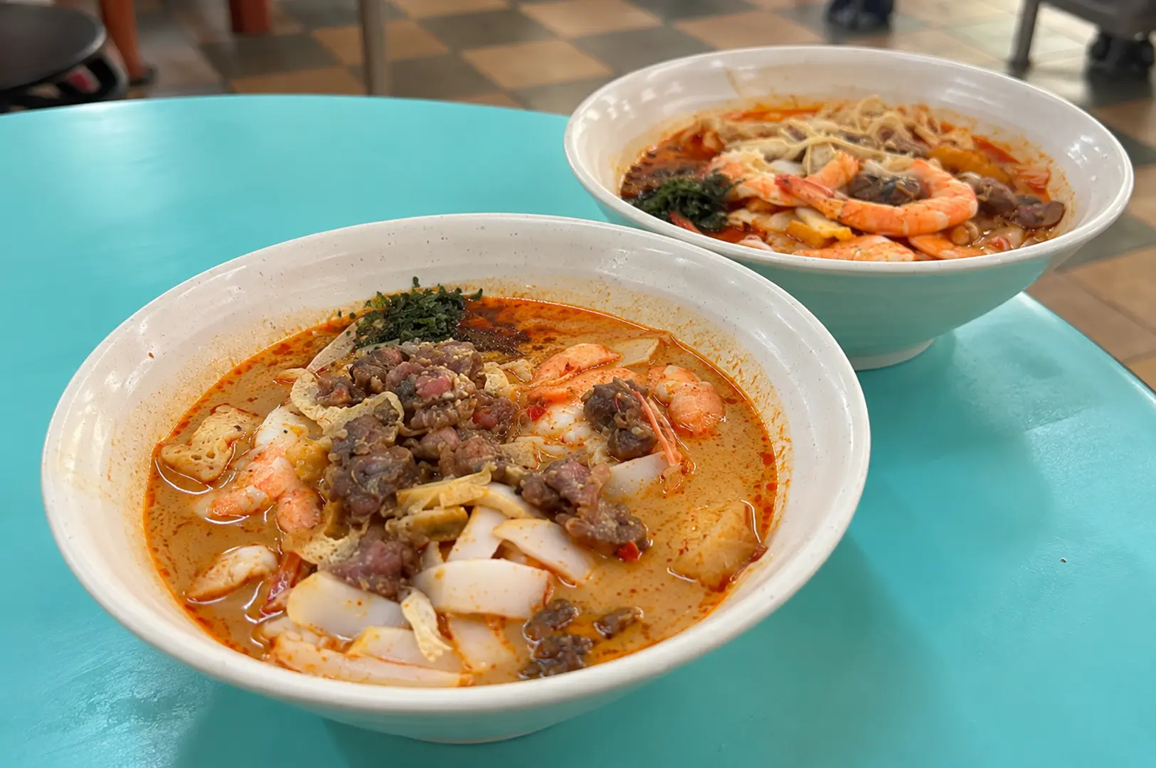 Two bowls of rich, creamy laksa served on a turquoise hawker-centre table, topped with prawns and other ingredients.