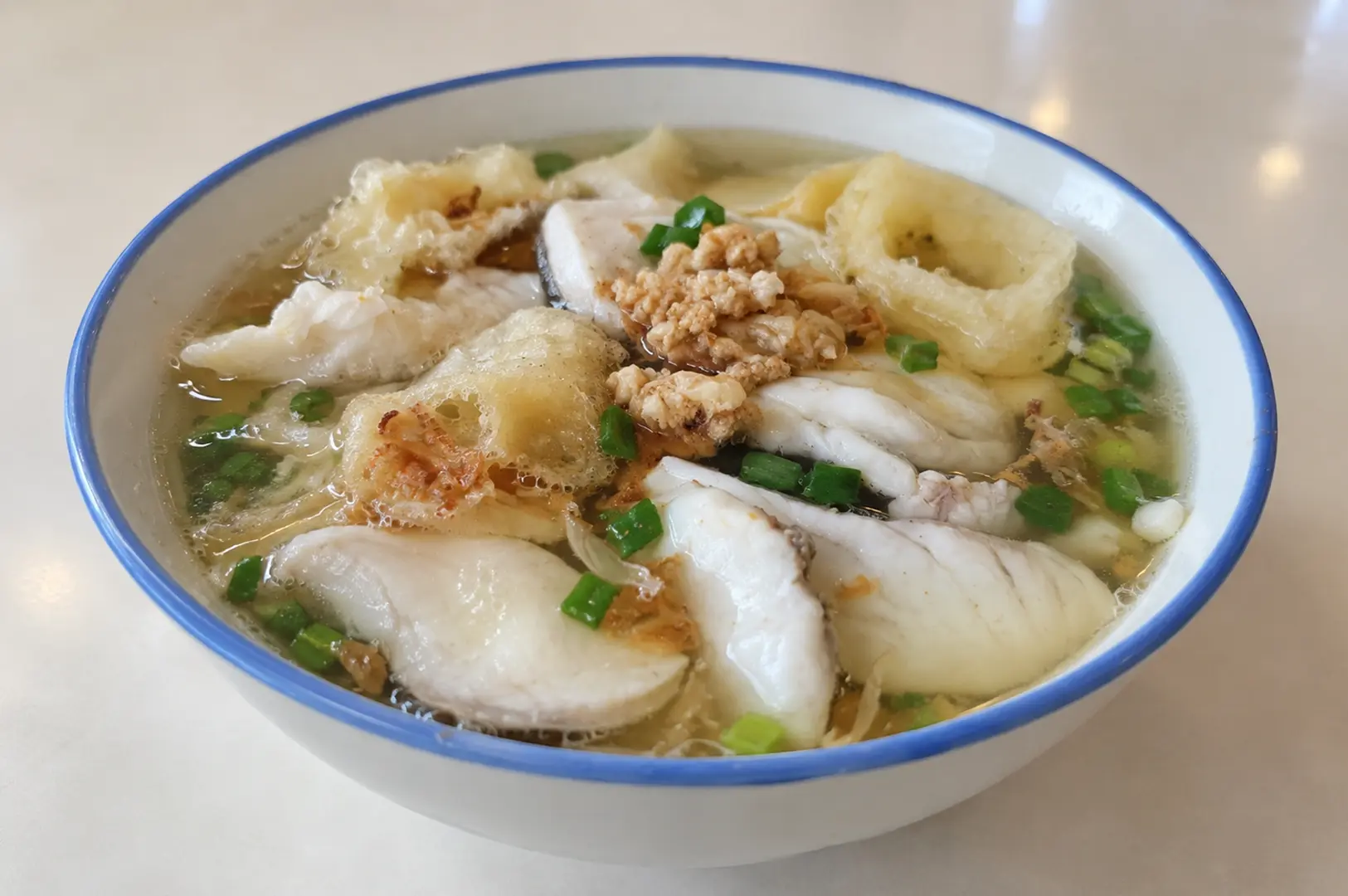 Bowl of fish soup with sliced fish, fried fish pieces, dumplings, and chopped green onions in clear broth.