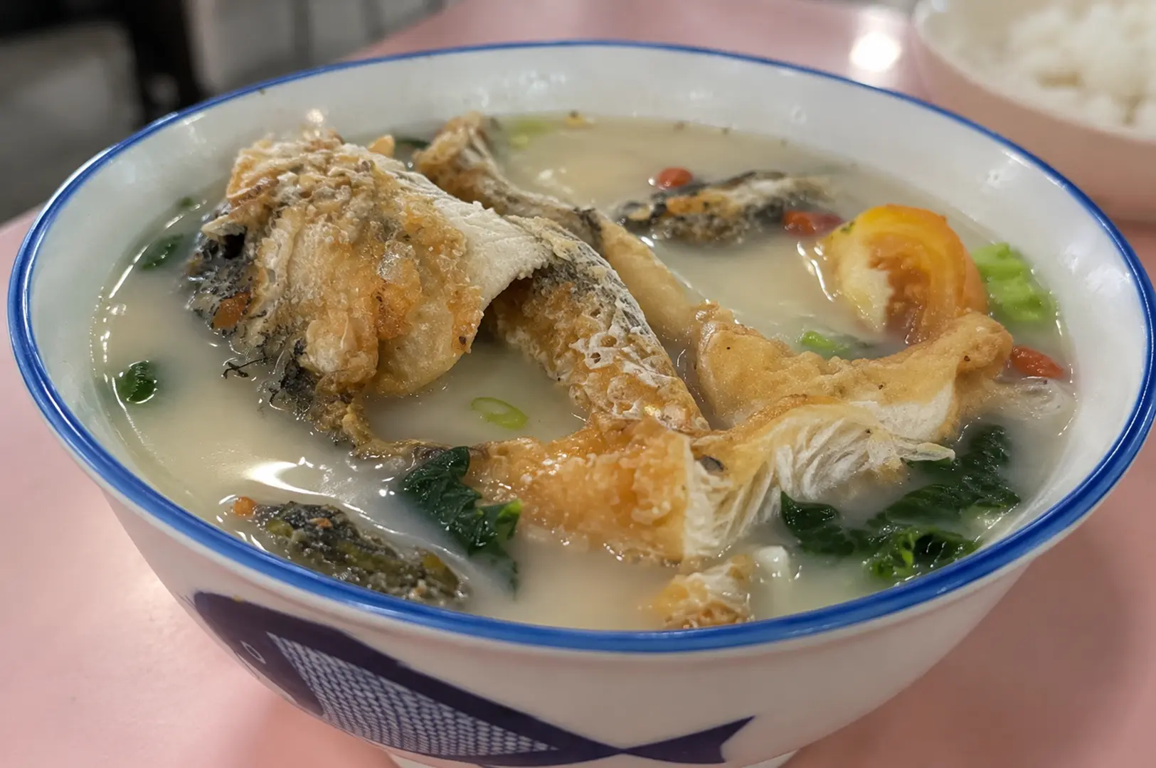 Bowl of fish soup with fried fish pieces, leafy greens, tomato, and vegetables in a light broth.