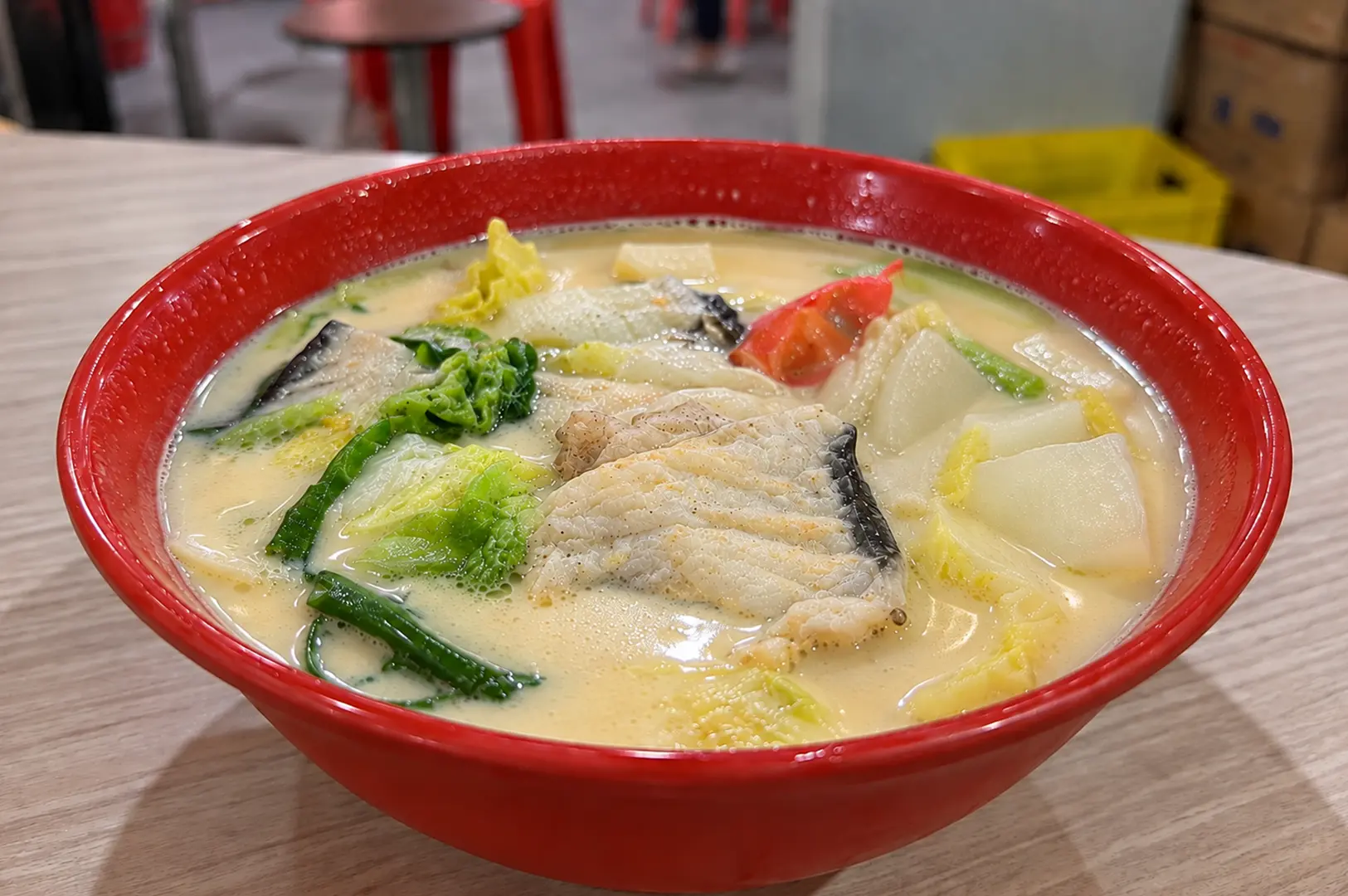 Creamy fish soup with sliced fish, leafy vegetables, and other fresh ingredients in a red bowl.