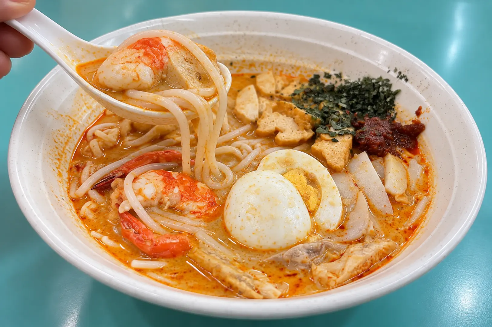 Close-up bowl of Singapore laksa with prawns, noodles, egg and tofu, with a spoon lifting noodles from the broth.