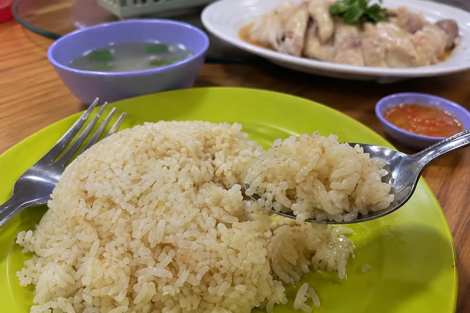 A spoon lifting glossy chicken rice from a bright green plate, with chilli sauce, clear soup, and sliced chicken in the background.