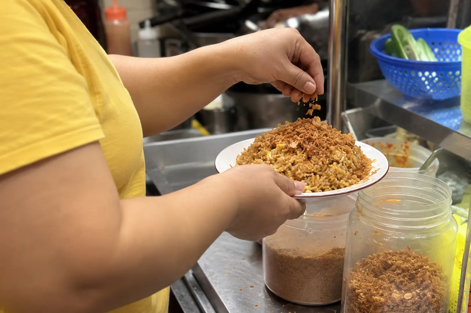 A street food vendor adds crispy fried shallots to a plate of fried rice at a hawker stall.