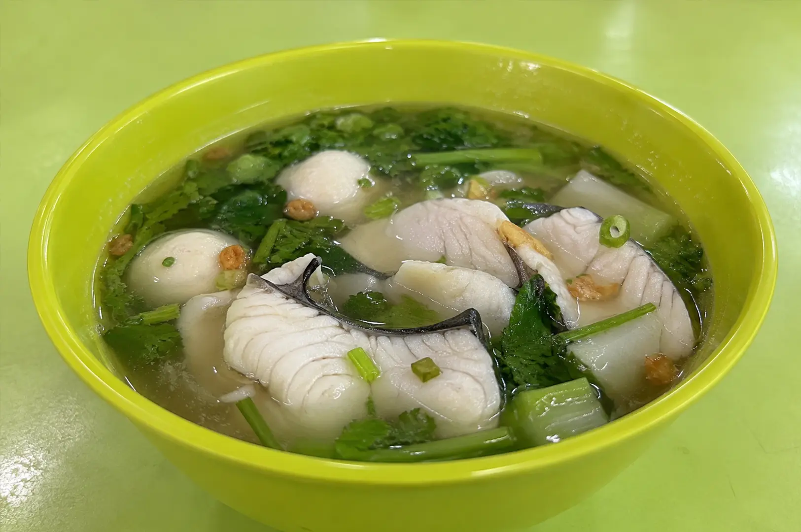 Yellow bowl of clear fish soup with sliced fish, fish balls, leafy greens, herbs, and crispy garlic toppings.