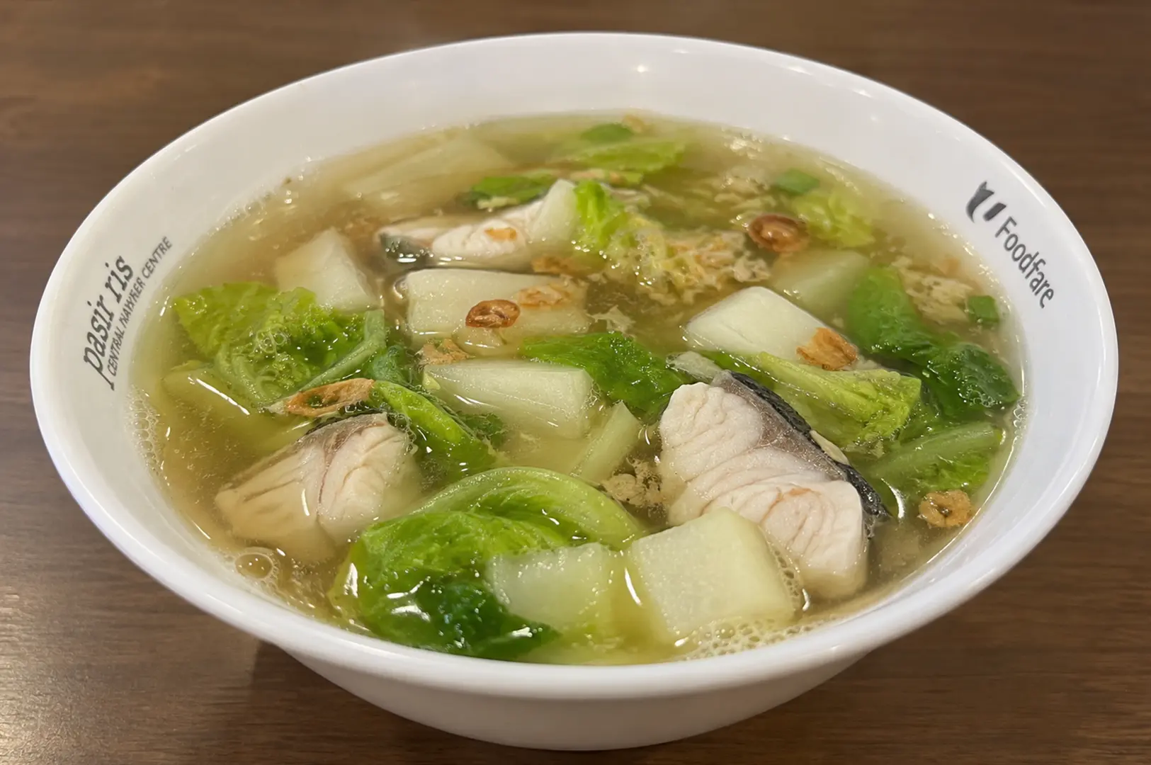 White bowl of clear fish soup with tender fish slices, cabbage, leafy vegetables, and crispy fried shallots.
