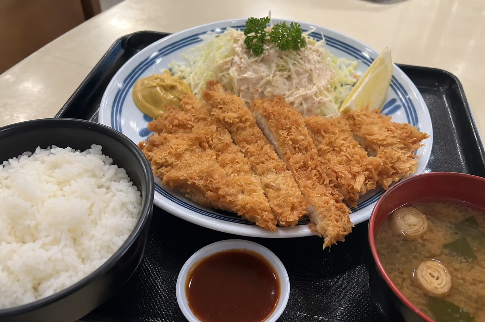 Generous serving of sliced golden pork katsu with steamed rice, shredded cabbage, dipping sauce, and miso soup on a black tray.