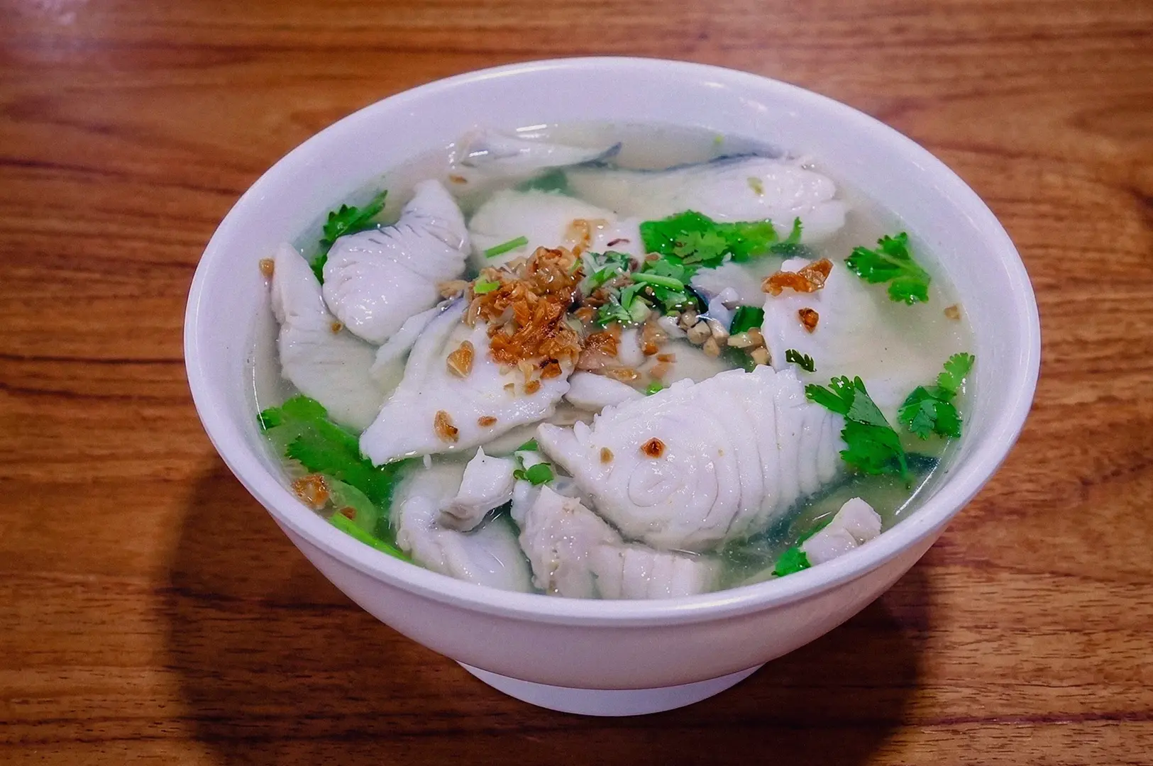 Bowl of clear fish soup topped with sliced fish, fresh cilantro, and crispy fried garlic on a wooden table.