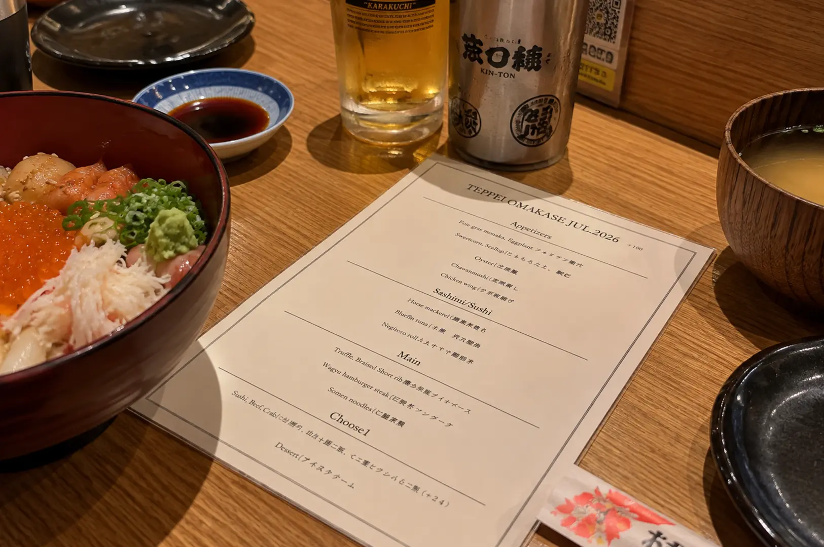 Teppei omakase dinner menu displayed beside a seafood bowl, miso soup, and drinks on the wooden counter.