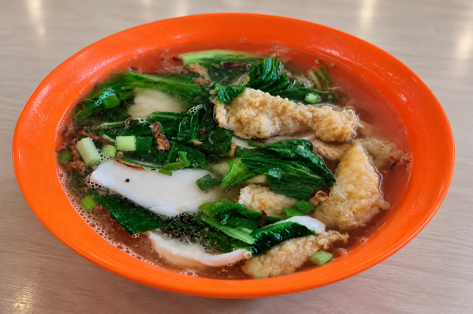 Orange bowl of fish soup filled with leafy greens, fish slices, and golden fried ingredients in clear broth.
